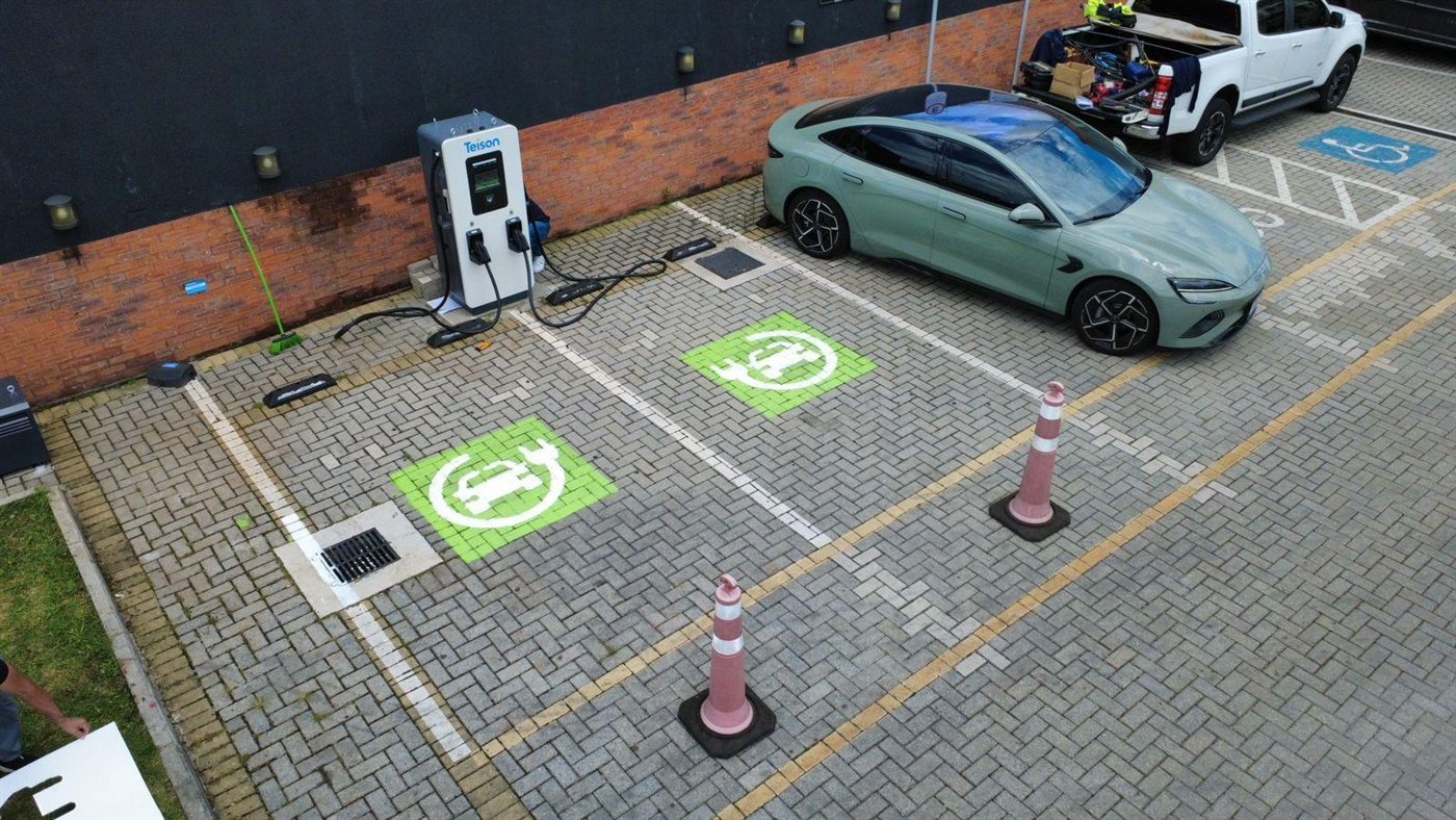 Vista aérea de um ponto de recarga com carregador rápido e um carro elétrico conectado, em vagas demarcadas para veículos elétricos